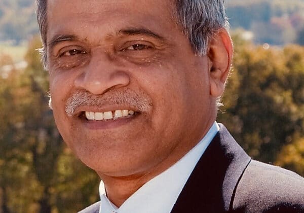 A man with short salt and pepper hair and a mustache smiles at the camera. He has medium-toned skin and is wearing a white collared shirt with a yellow tie and black dress coat. Autumn trees are blurred in the background.