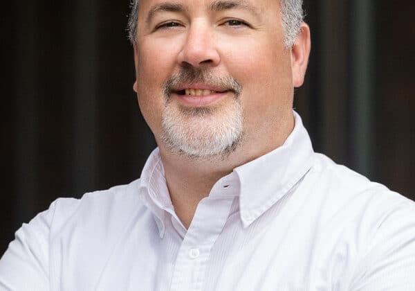 A man with salt and pepper hair at the temples, a goatee and mustache smiles at the camera. He has light-toned skin and is wearing a white collared shirt. The background is blurred.