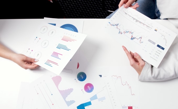 Hands of two people stretch across a white table, reviewing a sheaf of print-outs of market reports and charts.