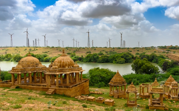 View of Bada Bagh, or Barabagh in the Indian state of Rajasthan. Decorative older buildings in the foreground and a river and large wind turbines in the background.