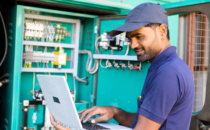 An engineer with medium-toned skin, wearing a blue polo shirt and cap is reading a laptop. In the background is an electrical system grid board.