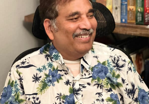 A man sits in an office chair, smiling at someone to his left. He has short dark hair, medium-toned skin, and is wearing a cream and blue floral print shirt. Shelves of books are in the background.