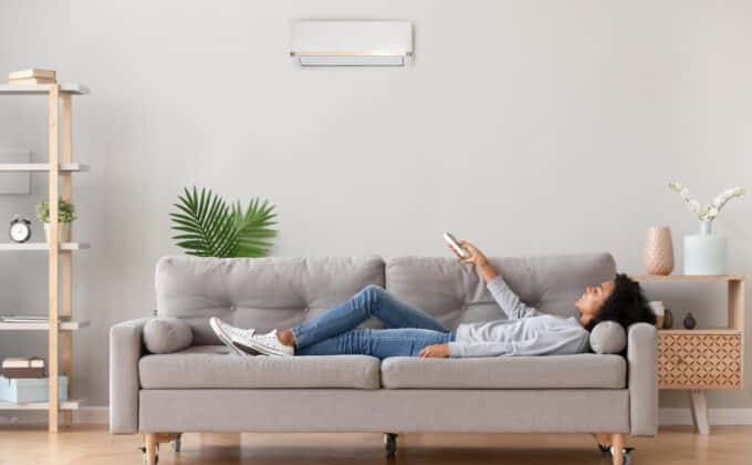 Relaxed woman in casual wear lying on sofa in living room at modern home holding heat pump remote control