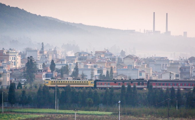 A coal mining town in CHina, with a train passing in the foreground and a refinery on the mountainside in the background. Emissions cloud the sky.