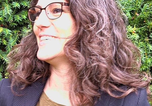 A woman with dark curly hair looks to the right of the camera. She is wearing a black blazer and a brown shirt. She has light-toned skin and dark framed glasses. Green hedges are in the background.