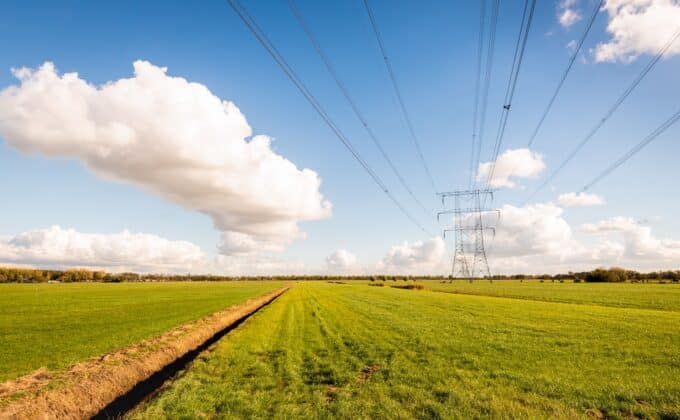 High voltage lines and power pylons in a Dutch agricultural landscape with large meadows. Diagonal in the image is a long ditch. It is a sunny day with a bright blue sky in the autumn season.