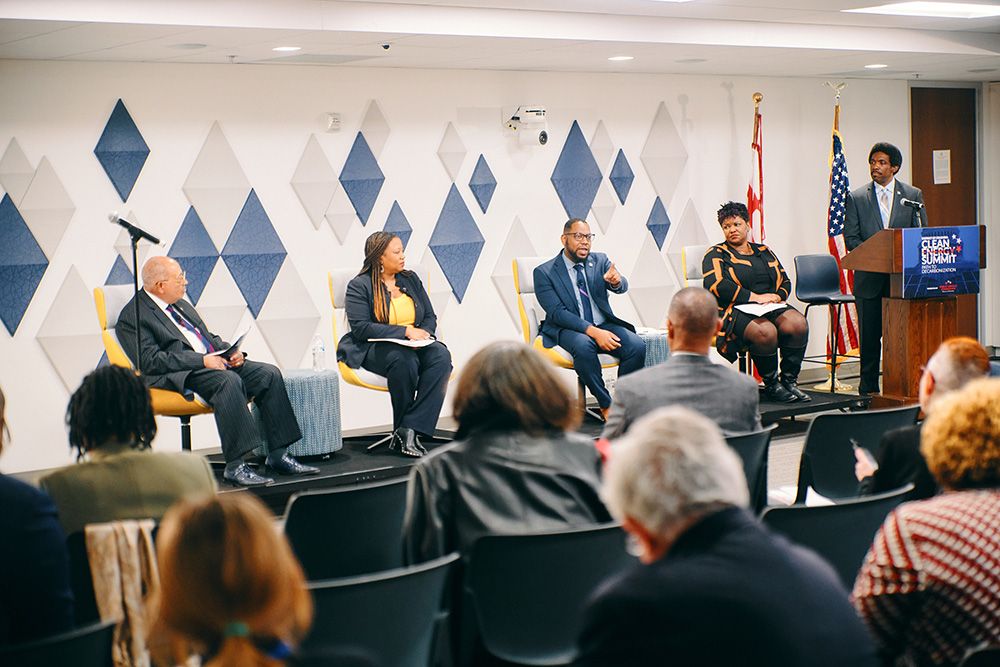 Four speakers sit on a panel for a Washington D.C. clean energy summit. A moderator stands at a podium to the far right. Credit: Public Service Commission of the District of Columbia