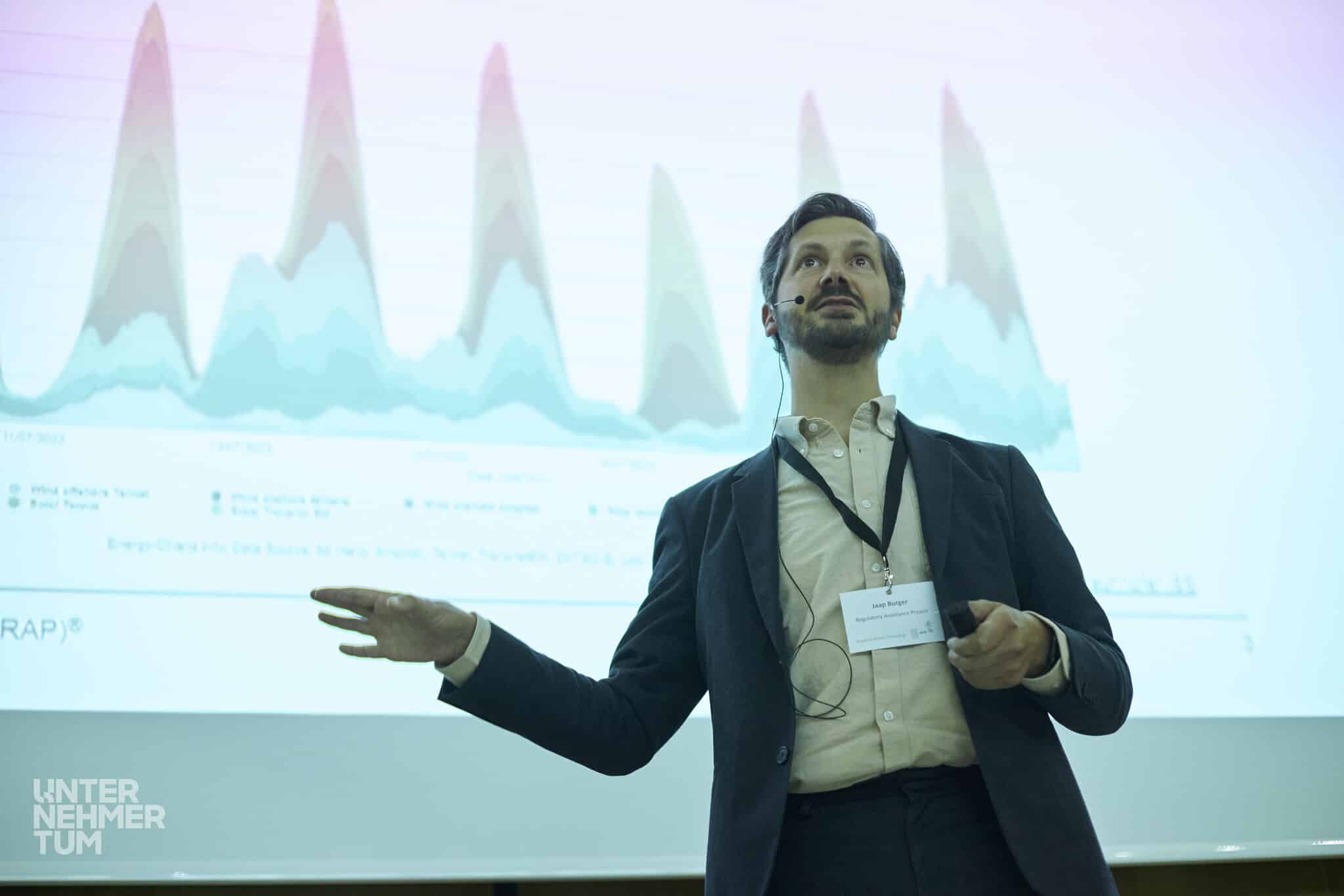 A man with dark hair and a full beard stands in front of a screen projected chart. He is wearing a suit and speaking to a crowd we cannot see.