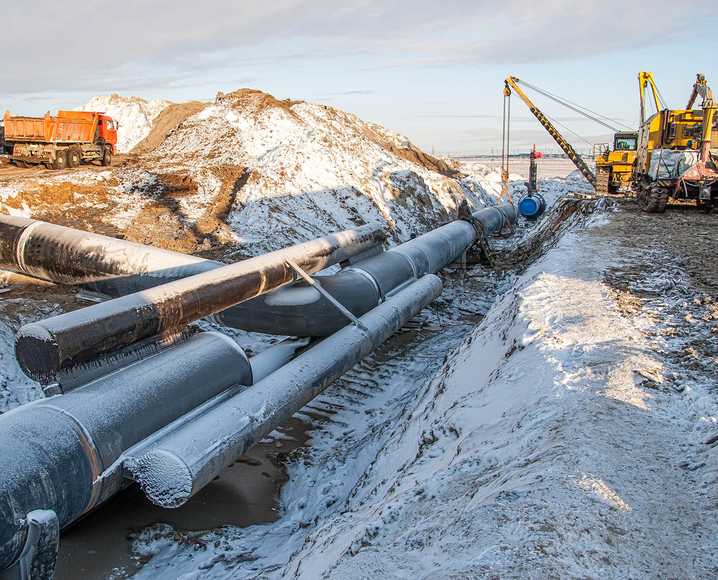 A trench has been dug into the ground and gas pipeline is being laid down. Construction equipment is to the right of frame. A dusting of snow covers the ground.