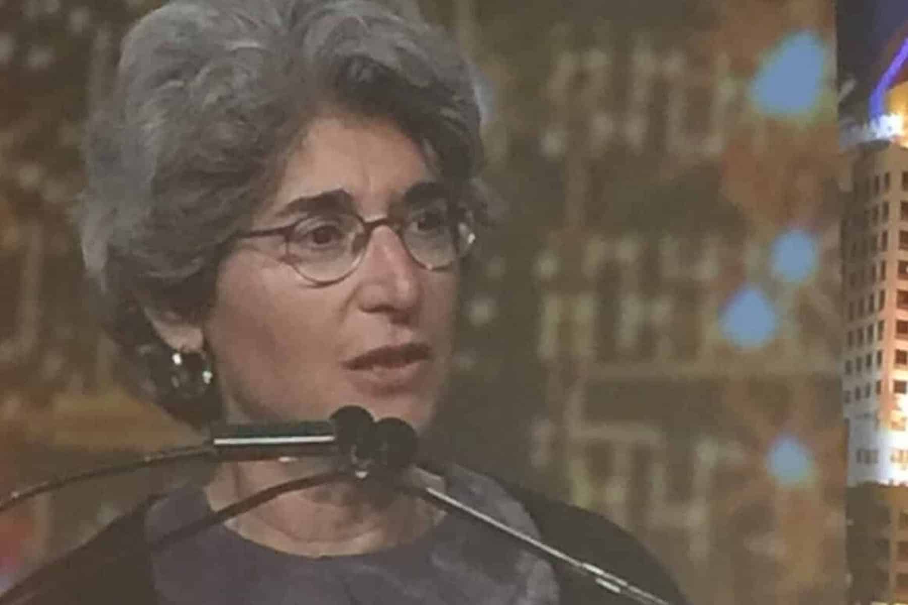 A woman with curly salt and pepper hair is standing at a podium, speaking into a microphone. She wears glasses and is being presented an award. Nancy Seidman receives AWMA award.