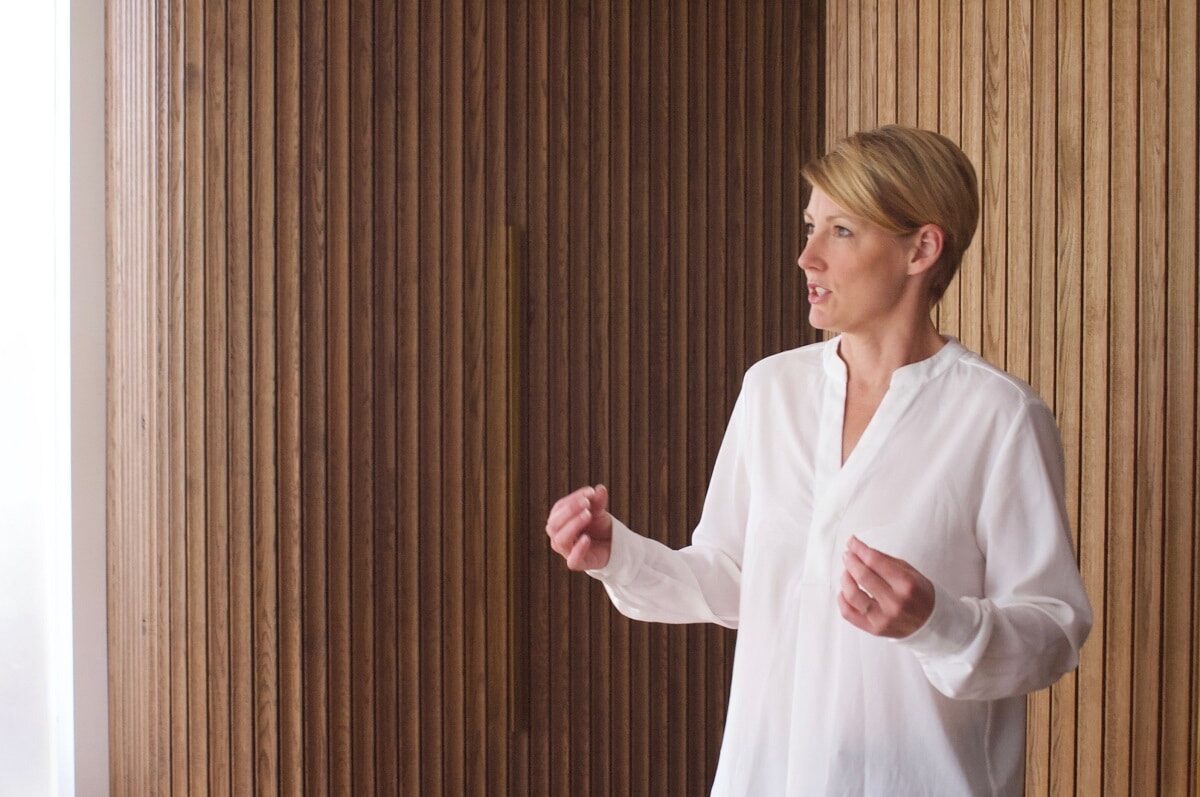 A woman with short cropped blond hair and wearing a loose flowing white blouse is in the middle of speaking. Behind her is a wood-paneled wall.