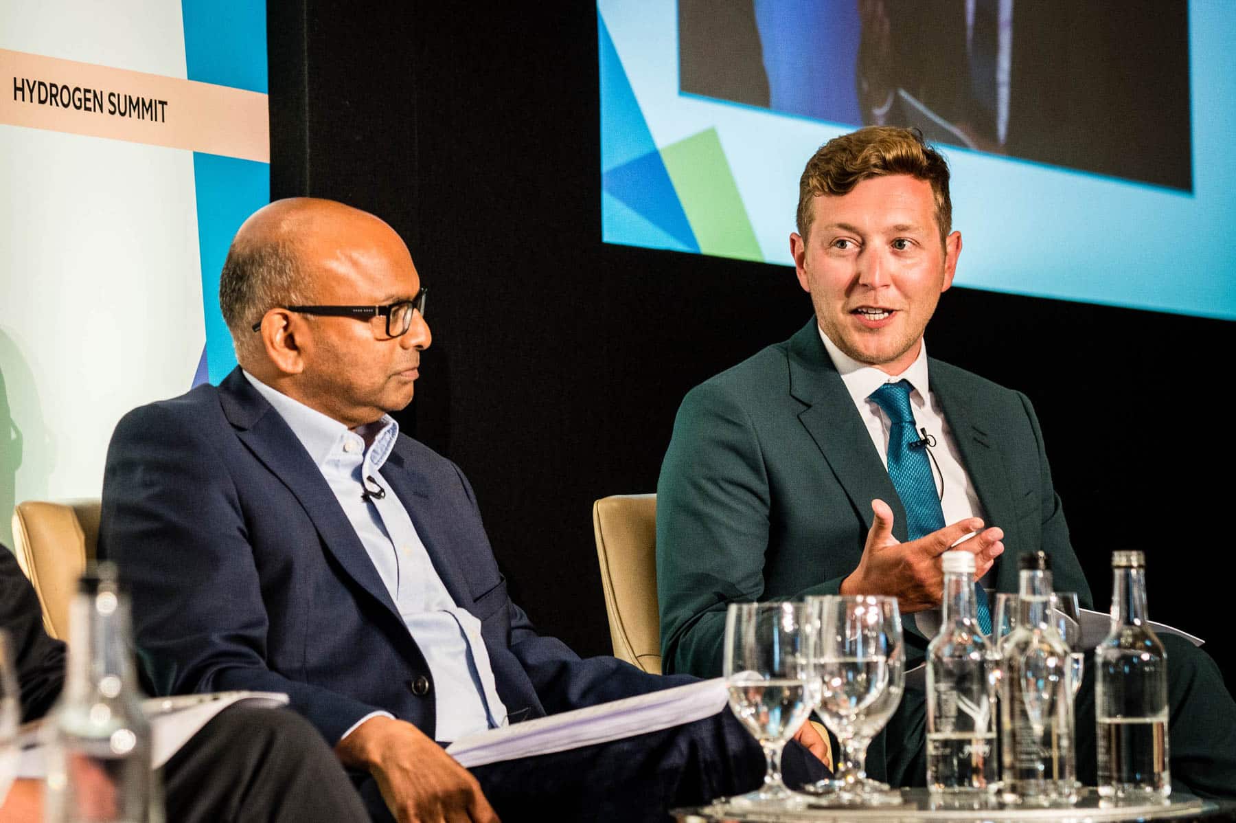 Two men sit on a stage in front of a screen projection. Both men are wearing suits and are panelists for an H2 event.