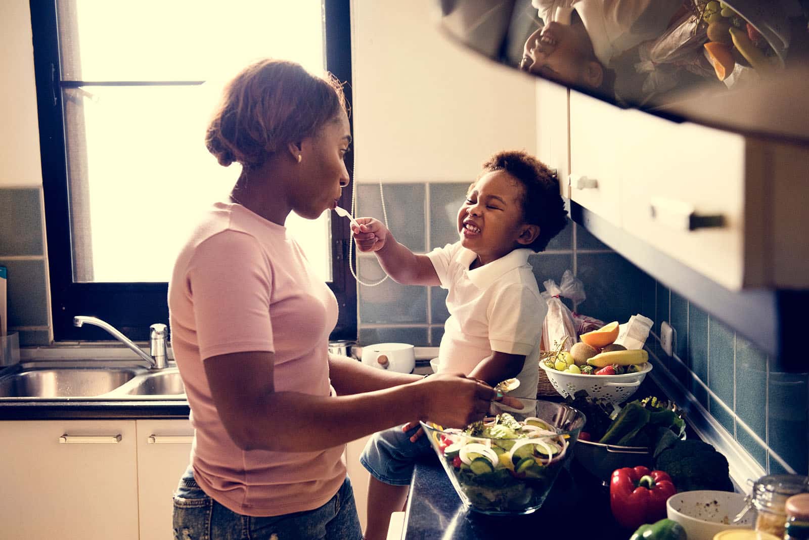 A woman with dark toned skin is preparing a salad in her kitchen, she is wering a pink t-shirt. On the counter next to her is a toddler, making a big smile and feeding her from a fork. 
