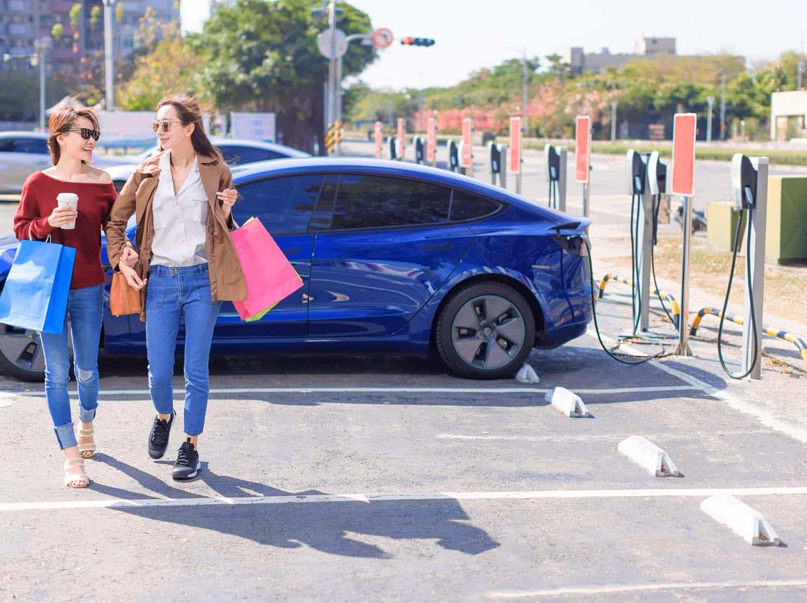 Happy young women standing on city parking near electric car, charging automobile battery from small city station, holding shopping bags and walking together.