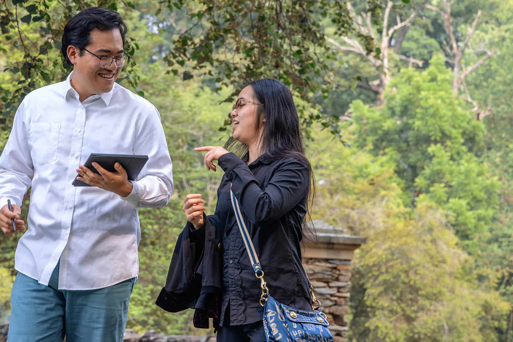 Two work colleagues discuss as they walk. The man has medium toned skin, black hair, and is wearing a white button down shirt. The woman has long, dark hair, is wearing a black blazer, and carries a satchel. He is holding a tablet. Green trees are in the background.