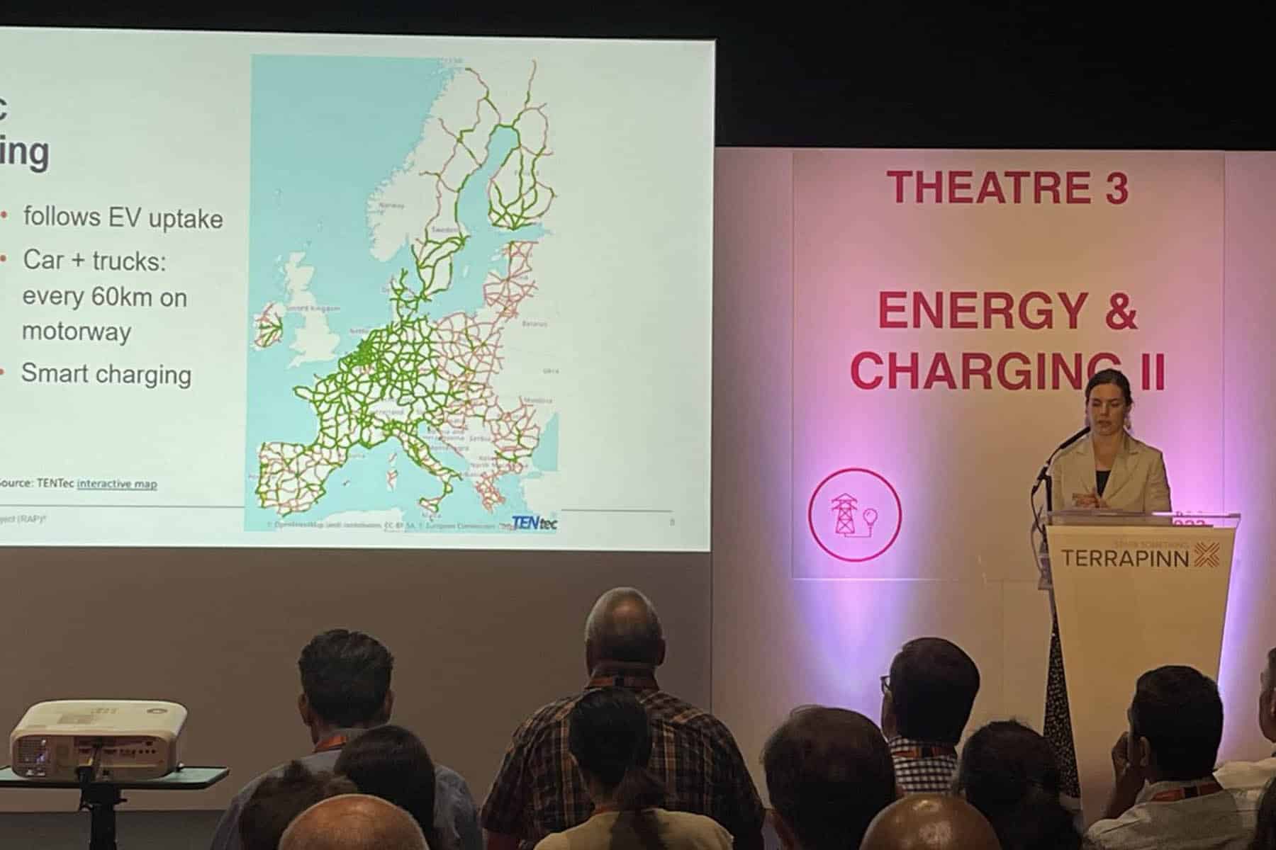 A woman with light toned skin stands are a podium in front of a group. To the left of frame is a screen projection of a map of Europe. In red letters on the wall behind her are the words "Energy & Charging II".