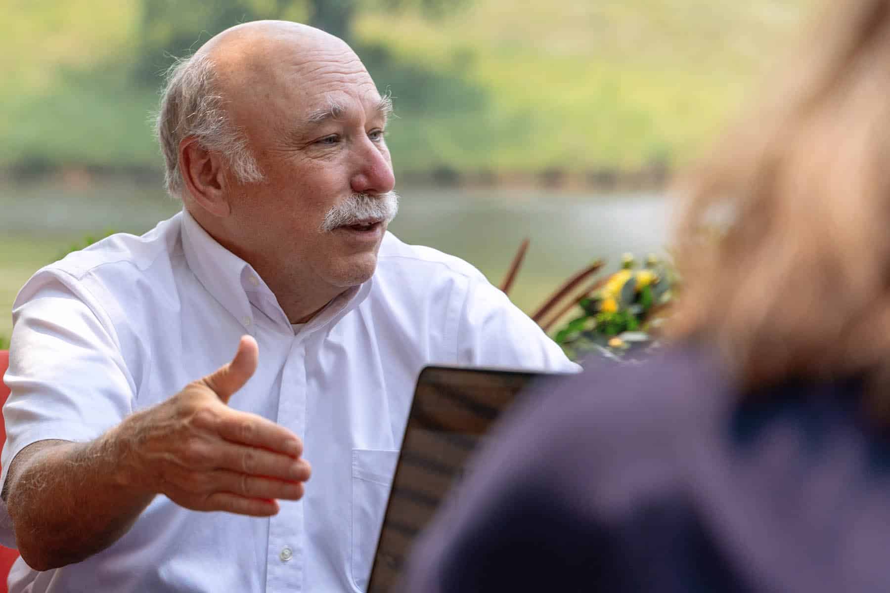 A man with white hair and a white mustache is sitting in front of a lake. He is wearing a short-sleeved white button down shirt and is speaking with a group off-camera.