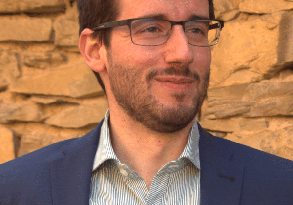 A man with dark curling hair and dark-framed glasses, smiles. He is wearing a light green shirt and a dark blue jacket.