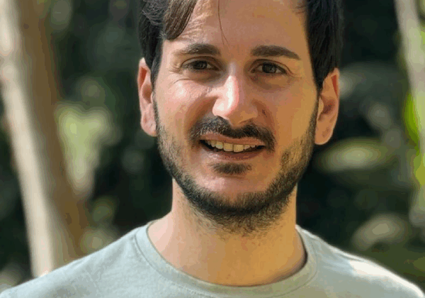 A man with light-toned skin smiles at the camera. He has dark hair and a full beard. He is wearing a light gray t-shirt.