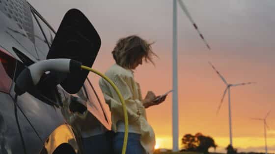 A woman wearing a yellow blouse and jeans stands outdoors at sunset, using her smartphone near an electric car charging by wind turbines.
