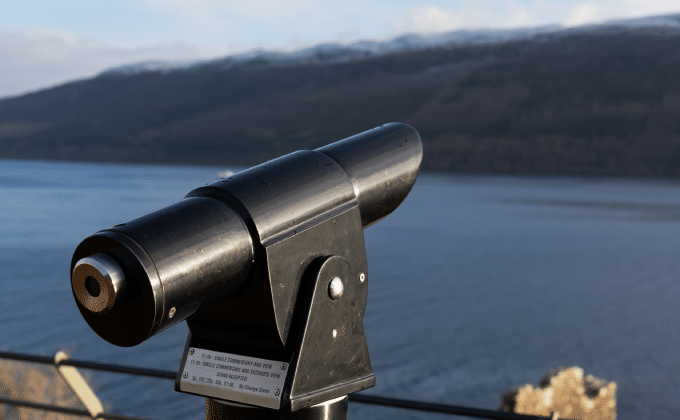 A viewing scope is perched at a railing, overlooking a waterway and treed hills in the distance.