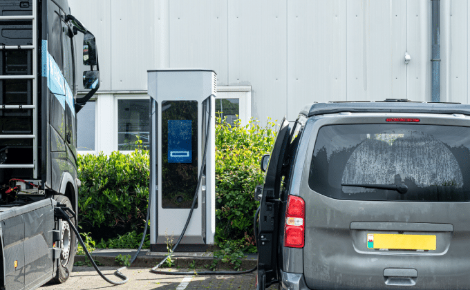 A heavy-duty truck and passenger van are parked on either side of a public EV charging station. A light-stone building is in the background.