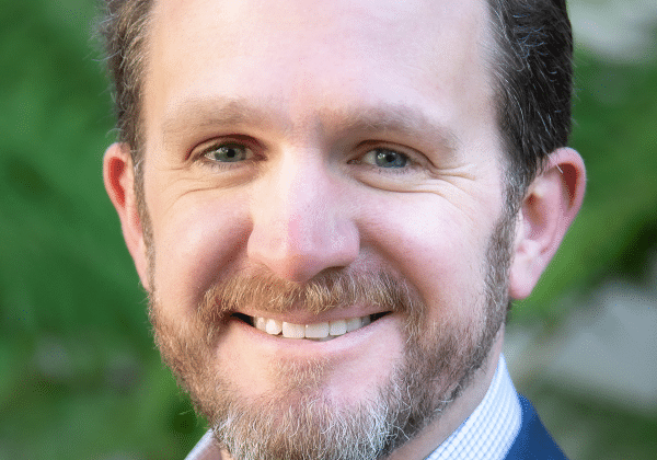 Man with dark, wavy hair and a full beard smiles at the camera. He has light-toned skin and is wearing a blue suit.